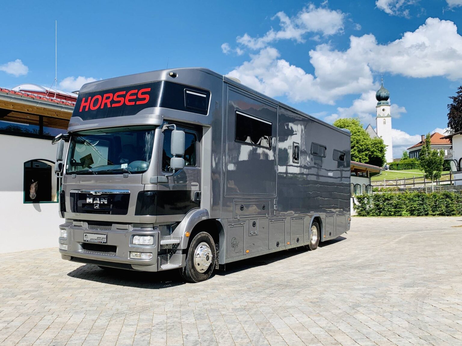 MAN-Pferdetransporter mit roter 'Horses'-Aufschrift vor Reitanlage mit Kirche